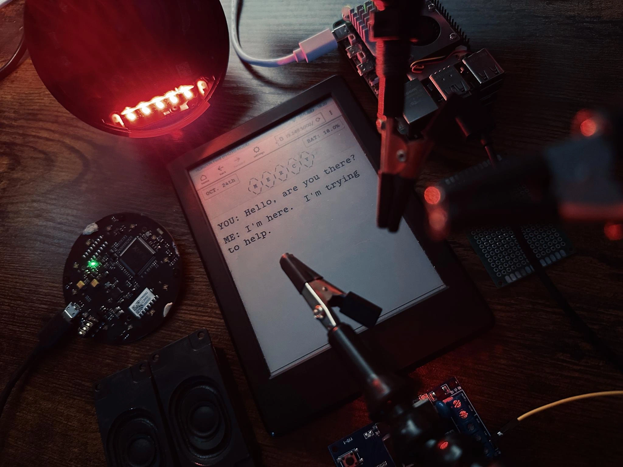 Close up of the Kindle device with other electronics around it.  the screen reads: Hello, are you there?  I'm here. I'm trying to help.