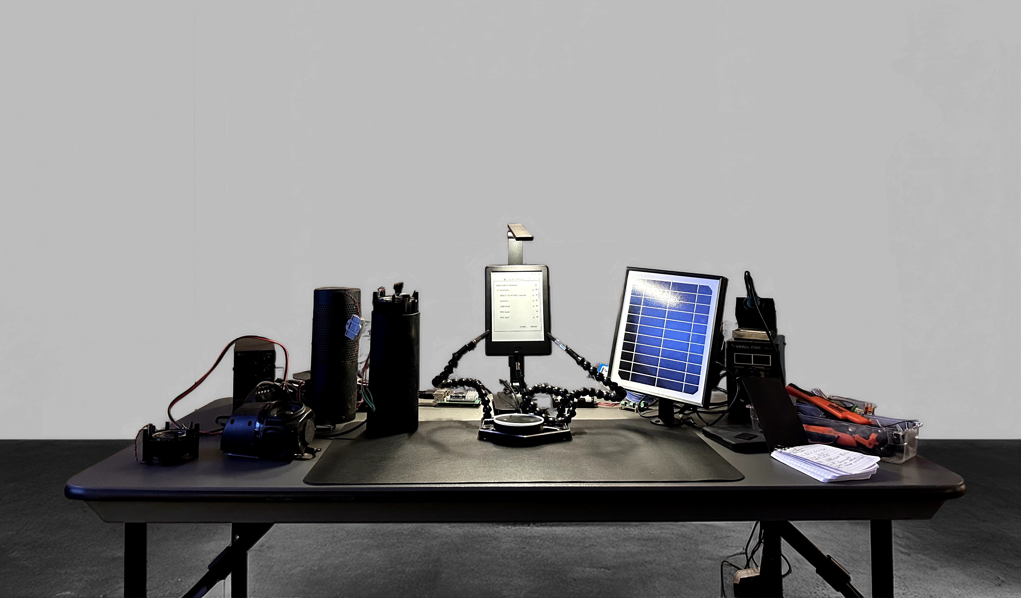 An image of The Hozion, a cluttered grey desk with black, hacked electronic devices surrounding a suspended Amazon Kindle.